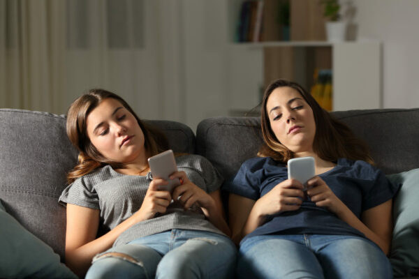 Twee pubermeisjes onderuit gezakt op de bank met beiden hun telefoon in de hand. Ze hebben alleen aandacht voor hun telefoon.