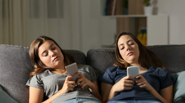 Twee pubermeisjes onderuit gezakt op de bank met beiden hun telefoon in de hand. Ze hebben alleen aandacht voor hun telefoon.