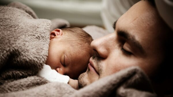 Baby slaapt bij vader onder een dekentje. Vader kijkt naar baby.